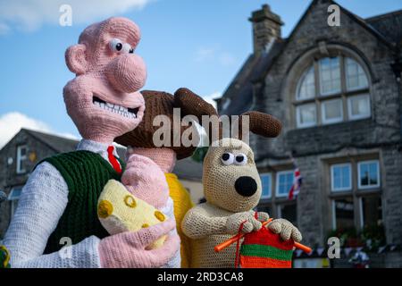 Hawes, North Yorkshire. 17. Juli 2023 Die Hawes Yarnbombers, eine lokale Gruppe extremer Stricker, enthüllt ihre neueste Erfindung in der Marktstadt Hawes im Herzen der Yorkshire Dales. Die überlebensgroßen gestrickten Figuren von Wallace und Gromit sowie andere Figuren aus der Serie nehmen mehrere Bänke ein und sehen Wallace mit einem großen Keil Wensleydale Käse, der sein Favorit ist und berühmt ist in der Wensleydale Creamery in Hawes. Spenden für die Kunstwerke gehen an den Yorkshire Air Ambulance Service. Kredit: Wayne HUTCHINSON/Alamy Live News Stockfoto