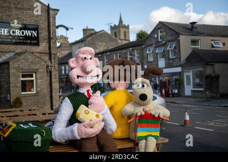 Hawes, North Yorkshire. 17. Juli 2023 Die Hawes Yarnbombers, eine lokale Gruppe extremer Stricker, enthüllt ihre neueste Erfindung in der Marktstadt Hawes im Herzen der Yorkshire Dales. Die überlebensgroßen gestrickten Figuren von Wallace und Gromit sowie andere Figuren aus der Serie nehmen mehrere Bänke ein und sehen Wallace mit einem großen Keil Wensleydale Käse, der sein Favorit ist und berühmt ist in der Wensleydale Creamery in Hawes. Spenden für die Kunstwerke gehen an den Yorkshire Air Ambulance Service. Kredit: Wayne HUTCHINSON/Alamy Live News Stockfoto