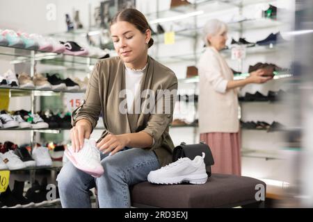Im Schuhgeschäft sitzt eine Frau auf einer Ottomane und probiert Turnschuhe an Stockfoto