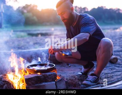 Ein brutaler Mann mit Bart kocht Essen in einer Bratpfanne auf einem Feuer in der Natur. Am Flussufer wird Speck gekocht. Camping, Sommer Stockfoto
