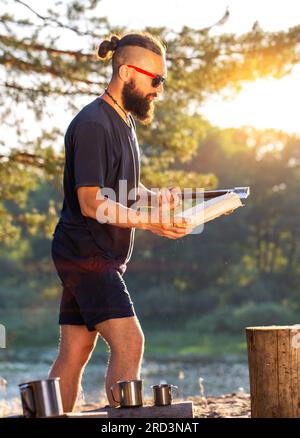 Ein eleganter, brutaler Mann mit Bart und schwarzer Brille hackt Holz auf einem Campingplatz, um ein Feuer anzünden. Outdoor-Aktivitäten im Sommer. Stockfoto