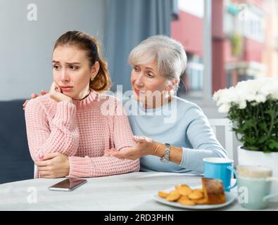 Vorsichtige ältere Mutter tröstet aufgebrachte Erwachsene Tochter am Tisch Stockfoto