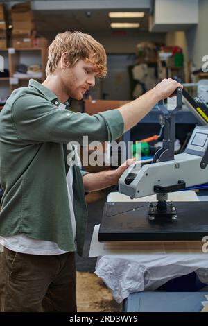Seitenansicht eines jungen rothaarigen Handwerkers, der mit einer Siebdruckmaschine und einem T-Shirt in einem verschwommenen Druckstudio im Hintergrund arbeitet, mit einem kundenorientierten kleinen Bu Stockfoto
