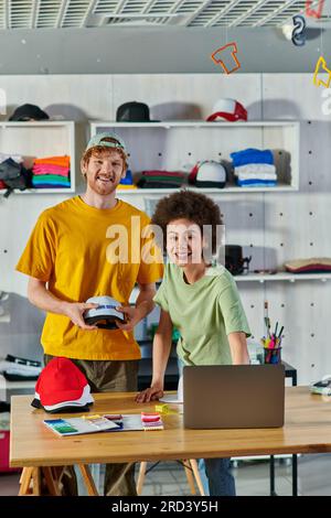 Fröhliche, junge, multiethnische Designer, die vor der Kamera arbeiten und dabei Snapbacks, Laptops und Stoffproben auf dem Tisch im Print Studio im Hintergrund verwenden, Stockfoto