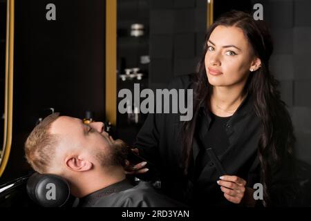 Hübsche junge Frau, männlicher Friseur, schneidet Männerbart mit Schere im Friseursalon Stockfoto