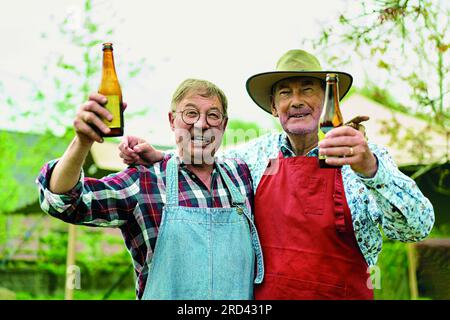 Zwei glückliche ältere Männer, tragen gemusterte Hemden und Barbecue-Schürzen, stoßen mit Weinflaschen in einem Landgarten an. Sie genießen ihre Zeit im Freien Stockfoto