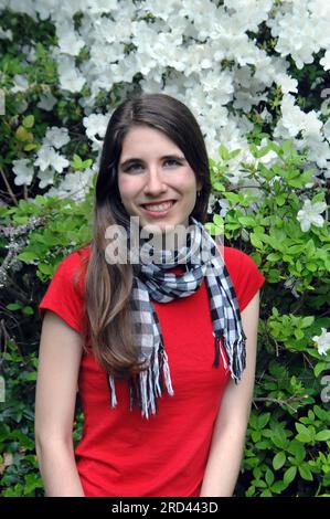 Teenager-Mädchen sitzt vor blühenden, weißen Azaleen. Sie lächelt und trägt ein rotes Hemd und einen schwarzen und weißen Schal mit Karomuster. Stockfoto