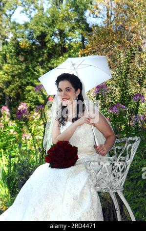 Die Braut posiert, sitzt auf einem weißen Metallstuhl. Sie hält einen weißen Sonnenschirm und lächelt. Ihr Strauß roter Rosen sitzt in ihrem Schoß. Stockfoto