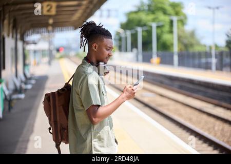 Junger Mann, Der Im Zug Zur Arbeit Pendelt, Steht Auf Der Plattform Und Schaut Auf Ein Mobiltelefon Für Reiseinformationen Oder Soziale Medien Stockfoto
