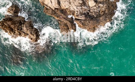 Blick aus der Vogelperspektive direkt über weißen Wellen, die auf rauen Felsen brechen, in einer rauen Umgebung an der Küste von Cornwall mit Platz zum Kopieren über Smaragdgrün Stockfoto