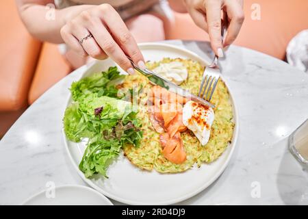 Frau schneidet Zucchini-Pfannkuchen mit Avocado, Käsesahne, Lachs und Ei. Gesundes Frühstück, Eiweiß. Restaurant-Gericht. Frühstück im Café bei Sonnenschein Stockfoto