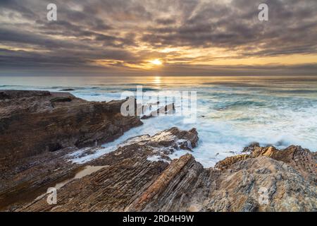 Felsige Küste bei Morro Bay, Kalifornien. Wellen brechen auf Felsen. Bewölkter Himmel mit sichtbarer Sonne, reflektiert vom Ozean. Stockfoto