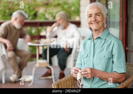 Fröhliche Seniorin mit weißem Haar, die in die Kamera schaut, während sie in einem Korbsessel auf der Terrasse des Altershauses sitzt und einen Strickschal trägt Stockfoto