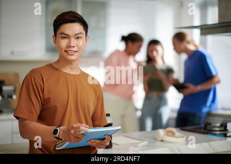 Porträt eines fröhlichen Studenten mit Lehrbuch, seine Freunde reden im Hintergrund Stockfoto