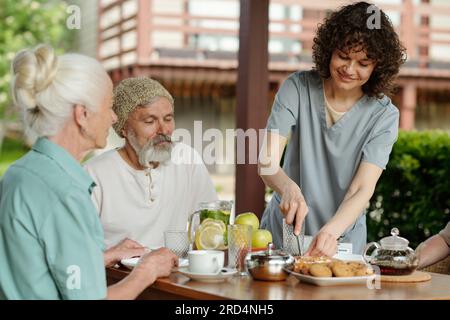 Junge, hübsche Pflegerin, die Kuchen oder Kuchen für eine Gruppe von Senioren schneidet, die zum Frühstück am Tisch sitzen und auf Tee mit Dessert warten Stockfoto