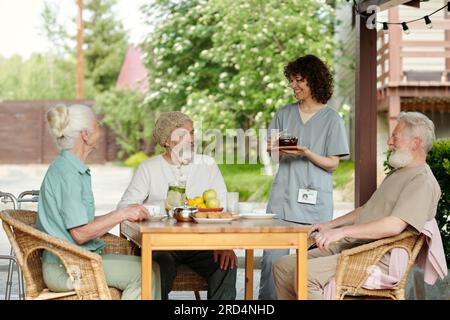 Eine glückliche junge Krankenschwester in Uniform brachte Teekanne mit Kräutertee für Patienten des Altershauses, die am Tisch mit Obst und einer Kanne Limonade versammelt waren Stockfoto