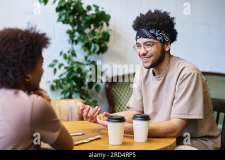 Glücklicher junger multiethnischer Mann in Freizeitkleidung, der mit seiner Freundin oder seinem Kollegen spricht, während er mit zwei Tassen Kaffee im Café am Tisch sitzt Stockfoto