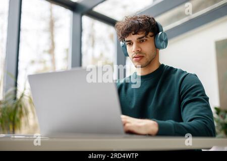 E-Learning-Konzept. Fokussierter europäer mit Kopfhörern, der sich ein Notebook ansieht, einen Online-Vortrag ansieht, im Klassenzimmer sitzt Stockfoto