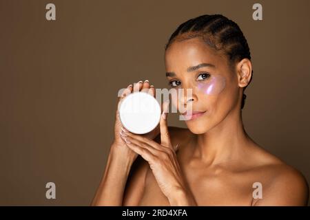 Attraktive schwarze Frau mittleren Alters mit Hydrogel-Augenflecken unter den Augen, die ein Glas hält und vor der Kamera zeigt, ein Modell Stockfoto