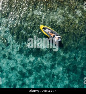 Ein einsames Weibchen in einem Strohhut lächelnd, entspannt im Kajak auf den türkisfarbenen Wellen des Adriatischen Meeres liegend. Luftaufnahme der Küste von oben. Exotisches c Stockfoto