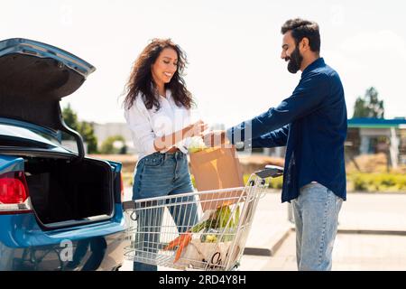 Glückliches Paar aus dem Nahen Osten, das Einkaufstüten in einen Kofferraum packt, arabischer Mann, der seiner Frau Papiertüte gibt Stockfoto