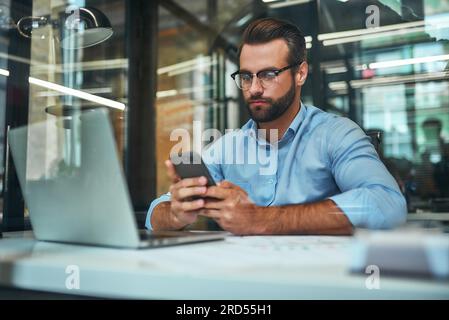 Wichtiger Anruf. Porträt eines jungen, fokussierten Geschäftsmanns mit Brille und formeller Kleidung, der sein Smartphone ansieht, während er im Büro sitzt Stockfoto