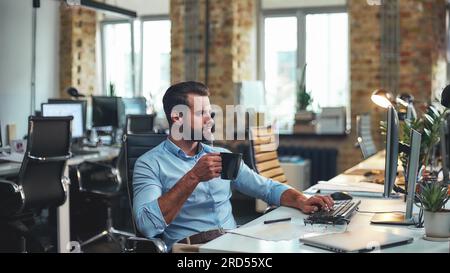 Frischer Kaffee. Seitenansicht eines jungen, bärtigen Geschäftsmanns in formeller Kleidung, der eine Tasse Kaffee hält und auf den Computer schaut, während er im modernen Büro sitzt Stockfoto