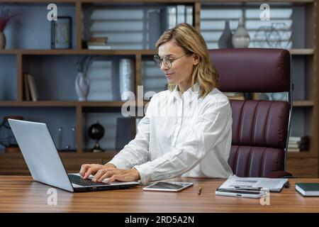 Erwachsen lächelnde Geschäftsfrau, die abends im Home Office arbeitet, Frau, die ein Notebook bei der Arbeit verwendet, Tastatur lächelt, Fernarbeit von zu Hause aus. Stockfoto