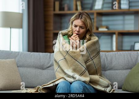 Alleinstehende, reife Frau, die zu Hause im Wohnzimmer auf dem Sofa sitzt, in eine Decke eingewickelt, um warm zu bleiben, fehlende und kaputte Heizung im Haus. Stockfoto