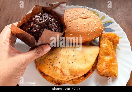 Auf einem Teller wird eine große Auswahl an Snacks ausgestellt. Eine Hand hält Schokoladenmuffin auf Holzhintergrund Stockfoto