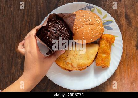 Auf einem Teller wird eine große Auswahl an Snacks ausgestellt. Eine Hand hält Schokoladenmuffin auf Holzhintergrund Stockfoto