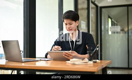 Ein fokussierter und seriöser asiatischer Geschäftsmann oder männlicher Büroangestellter, der sein Projekt plant und an seiner Arbeit auf seinem Tablet an seinem Schreibtisch arbeitet. Stockfoto