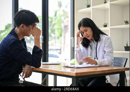 Eine gestresste und ernsthafte asiatische Ärztin hat ein ernstes Treffen mit einer Patientin in ihrem Untersuchungsraum in einem Krankenhaus. Stockfoto