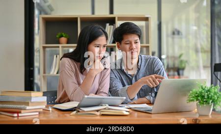 Zwei seriöse und fokussierte asiatische Studenten schauen auf einen Laptop-Bildschirm und diskutieren über ihre Arbeit, während sie zusammen in einer Campus-Bibliothek sitzen. Bildung Stockfoto