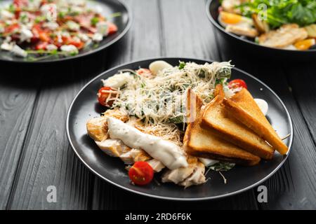 Verschiedene Salate auf einem Holztisch in einem Restaurant. Klassischer caesar-Salat, gebackene Paprika mit Fetakäse und Rucola-Salat auf dunklem Hintergrund, Top Stockfoto