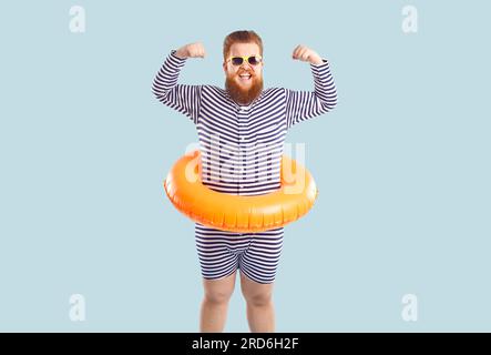 Der lustige, bärtige, fette Mann mit aufblasbarem Kreis zum Schwimmen in der Hüfte zeigt seinen Bizeps. Stockfoto