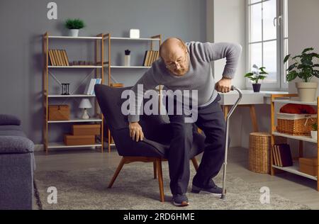 Ein Rentner mit schmerzenden Gelenken benutzt einen Gehstock, um vom Sessel aufzustehen Stockfoto