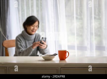 Unscharfe Frau, die lächelt und Smartphone liest. Selektiver Fokus auf eine Kaffeetasse und dunkle Schokolade auf einem weißen Tisch.