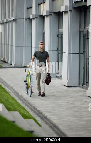 Bild des seriösen, konzentrierten, jungen, gutaussehenden Geschäftsmanns in eleganter Freizeitkleidung, der mit dem Fahrrad auf der Straße spaziert Stockfoto