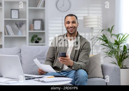 Porträt eines jungen afroamerikanischen Geschäftsmanns, der von zu Hause aus arbeitet. Er sitzt auf dem Sofa vor dem Laptop und hält ein Telefon und Dokumente in seinen Händen. Sie lächelte in die Kamera. Stockfoto