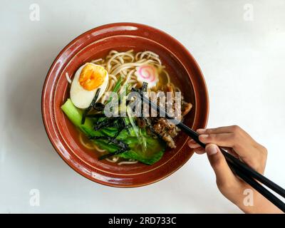 Hand verwendet Essstäbchen, um leckere japanische Ramen-Suppe in einer Schüssel auf einem weißen Tisch zu sammeln. Stockfoto