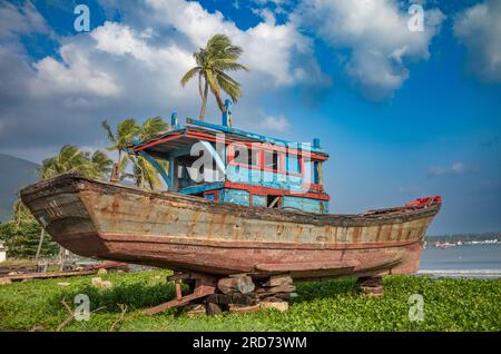 Ein zerschmettertes und unseetaugliches blaues und rotes Fischerboot aus Holz, das sich auf Häuserblocks neben dem Pham Van Dong Strand in Son Tra, Danang, Vietnam, befindet. Stockfoto
