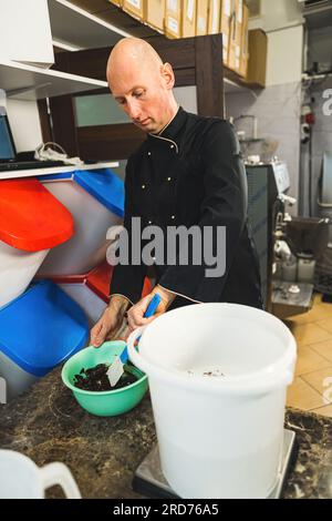 Vertikaler Schuss eines Konditorkochs in schwarzem Uniform, der Teig mit Spatel mischt. Kahlweißer Küchenarbeiter, der Eiscreme in der Industrieküche zubereitet. Hochwertiges Foto Stockfoto