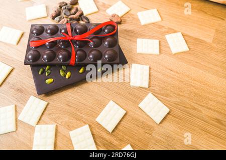 Zusammensetzung großer dunkler Schokoladenriegel mit kleinen, weißen Cholocatriegeln auf Holztisch mit rotem Band gebunden. Dunkle Schokolade mit Stücken, die wie Globen geformt sind. Dunkle Schokolade mit Pistazien. Hochwertiges Foto Stockfoto