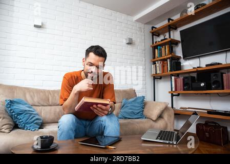 Ein lächelnder Millennials-Mann verbringt einen gemütlichen Abend im Wohnzimmer, sitzt auf der bequemen Couch und liest Lieblingspapierbuch, trinkt heißen Tee. Junger Leser hat Spaß enjo Stockfoto