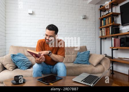 Ein lächelnder Millennials-Mann verbringt einen gemütlichen Abend im Wohnzimmer, sitzt auf der bequemen Couch und liest Lieblingspapierbuch, trinkt heißen Tee. Junger Leser hat Spaß enjo Stockfoto