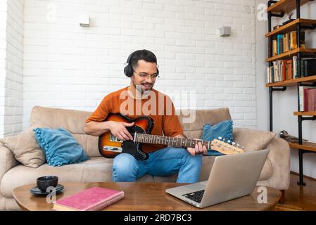 Junger Musiklehrer, der Schülern Gitarrenunterricht gibt, online auf einem Laptop von zu Hause aus. E-Gitarre mit dem Online-Kurs und spielen lernen Stockfoto