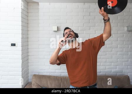 Junger glücklicher Mann, der zu Hause singt und tanzt und Musik auf dem schnurlosen Headset hört, auf seinem Telefon singt und LP-Platte in der Hand hält. Hipster-Musikliebhaber Stockfoto