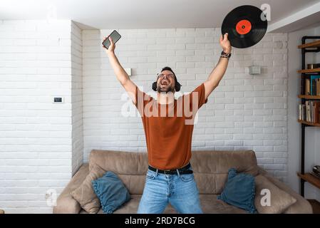 Junger glücklicher Mann, der zu Hause singt und tanzt und Musik auf dem schnurlosen Headset hört, auf seinem Telefon singt und LP-Platte in der Hand hält. Hipster-Musikliebhaber Stockfoto