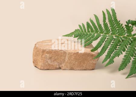 Steinernes Podium zur Beförderung auf neutralem beigefarbenem Hintergrund. Sockel aus Naturstein. Grüne Palmenfarnblätter. Modell von Schönheitsprodukten. Kosmetik, Seifenstand. Stockfoto